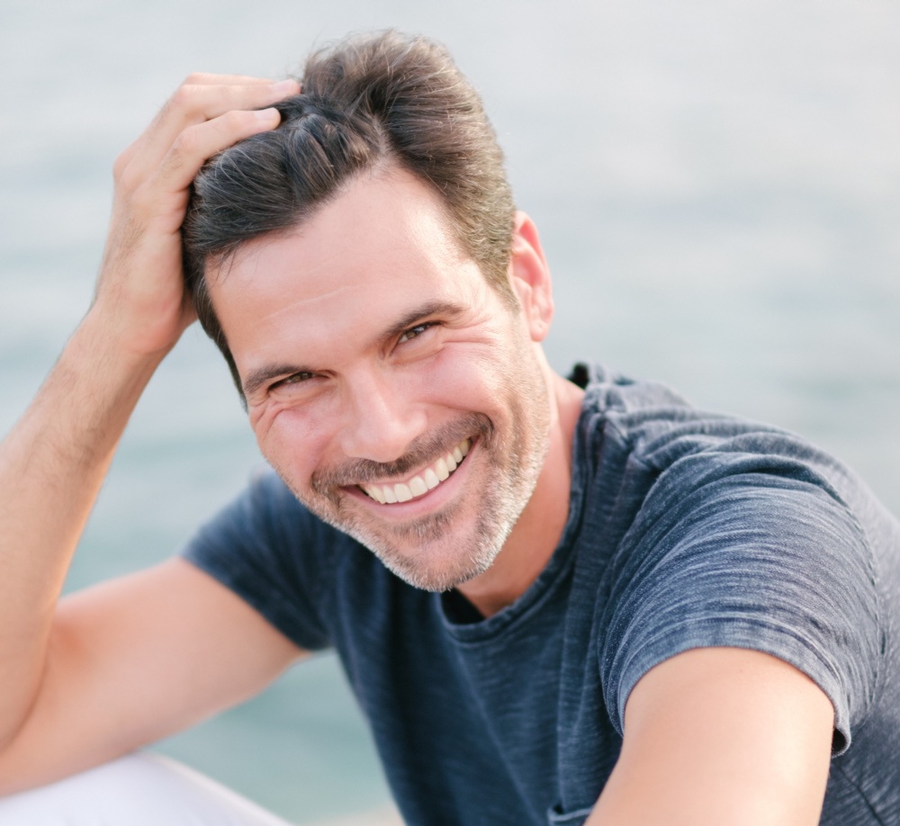 Delighted adult bearded male tourist in white pants and t shirt leaning on hand and smiling at camera while sitting on rocky coast near sea on sunny day
